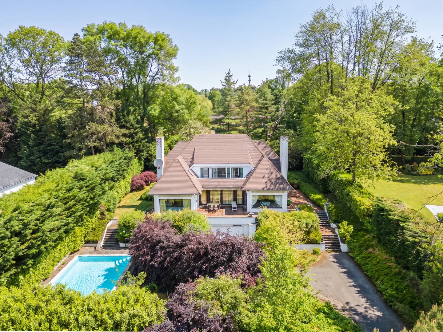 Aerial view of a white house with a brown roof, surrounded by green trees and a blue pool.