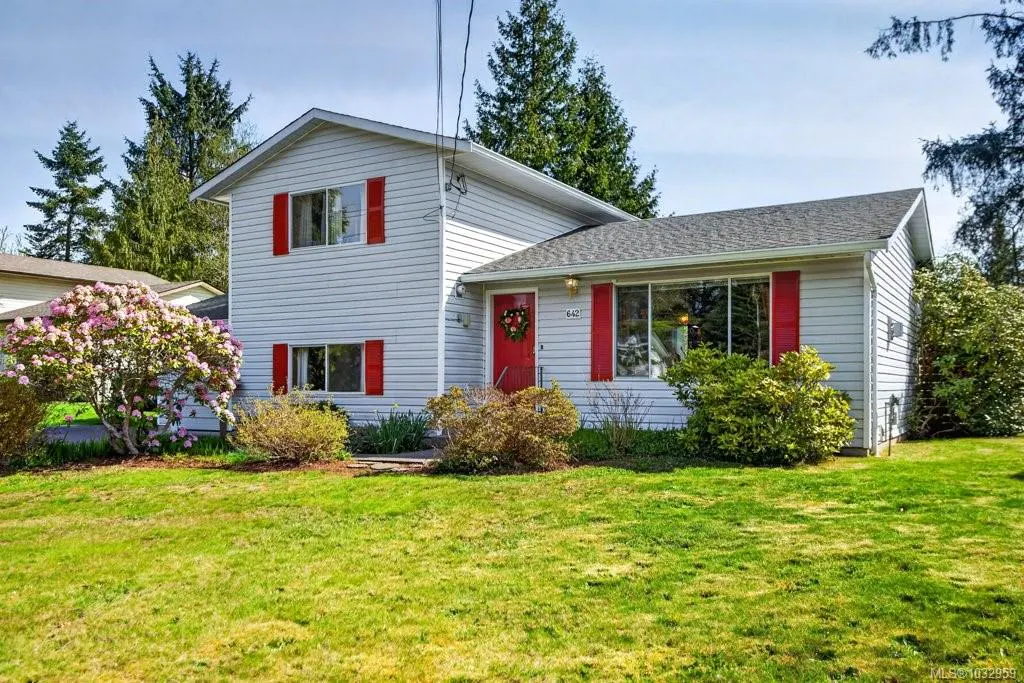 Two-story house with white siding, red shutters and door, and a green lawn. A flowering bush is on the left.