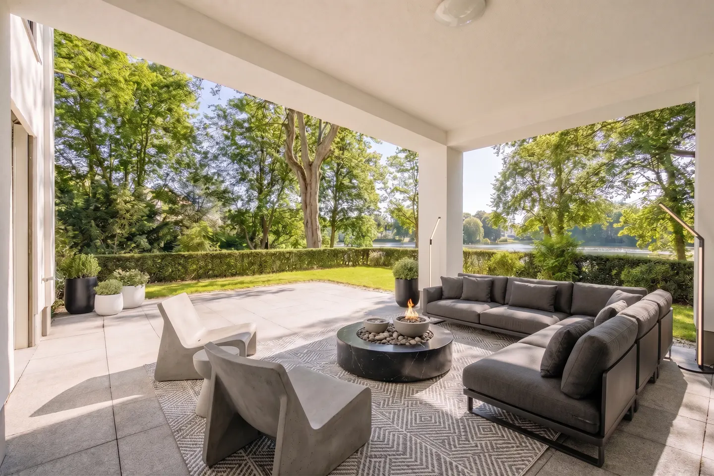 Outdoor patio with gray sofa, chairs, and fire pit on a patterned rug. Green trees and a lake are in the background.