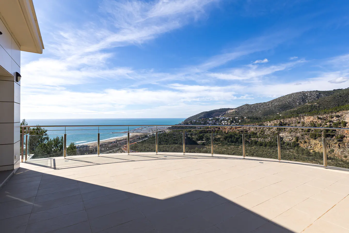 Balcony view of the ocean and mountains. Beige tile floor, glass railing, blue sky with clouds.