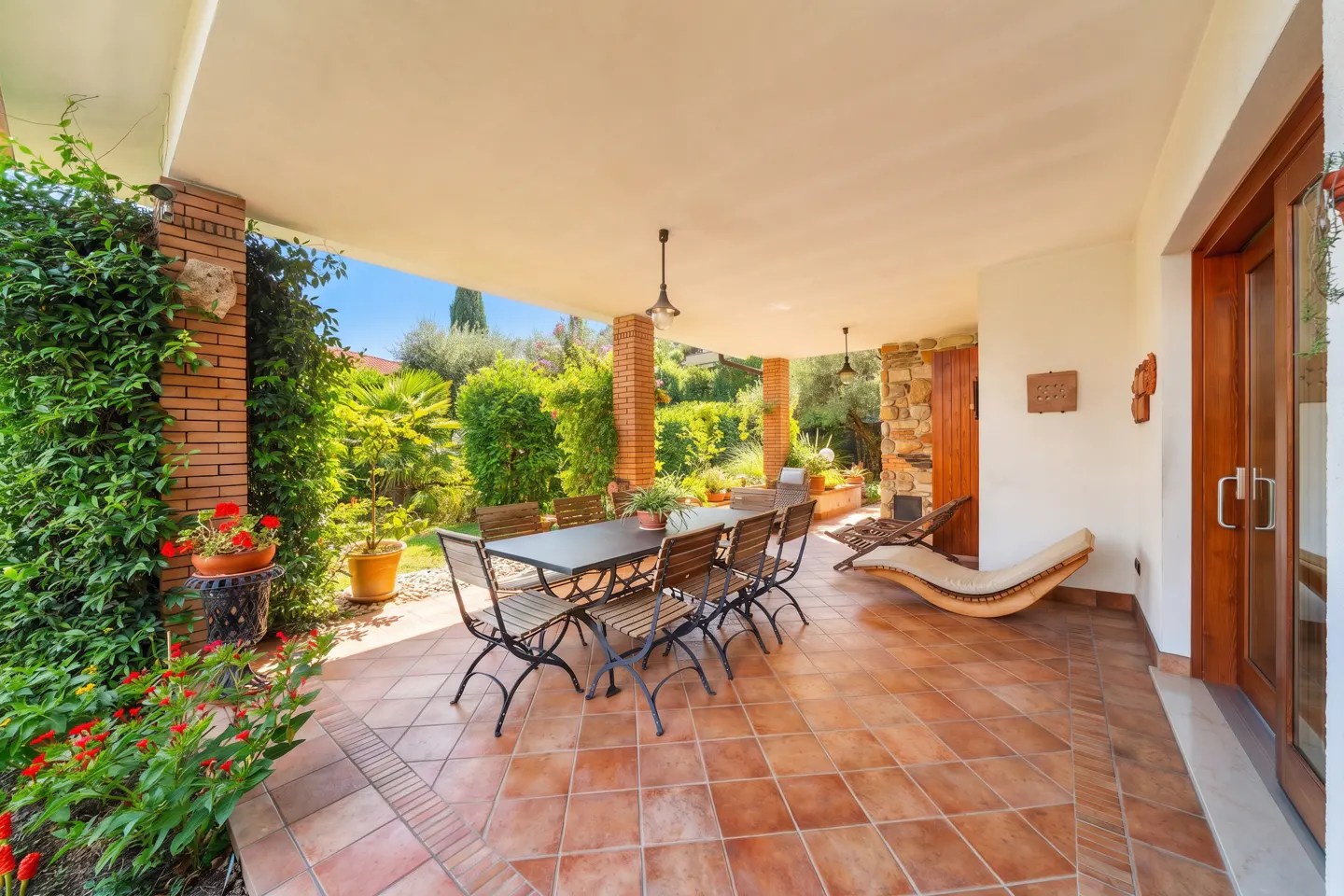 Covered patio with a dining table, chairs, and a lounge chair. Brick columns and lush greenery surround the space.