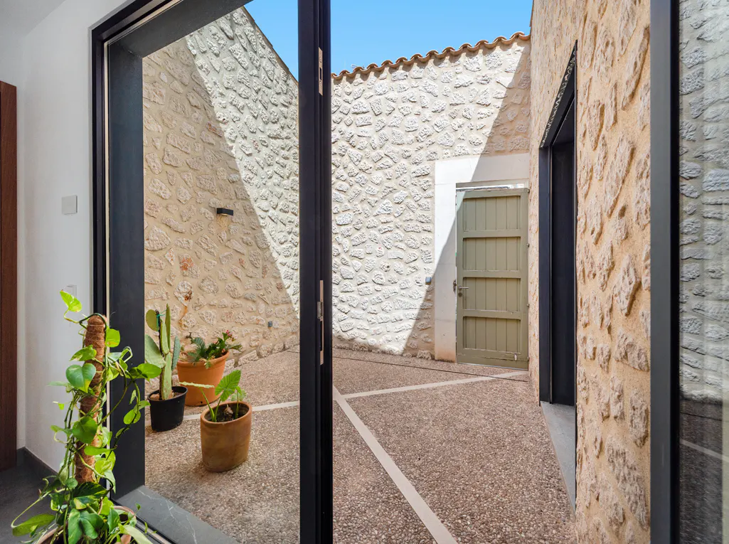 View through a black-framed glass door to a stone courtyard with potted plants and a light green wooden gate.