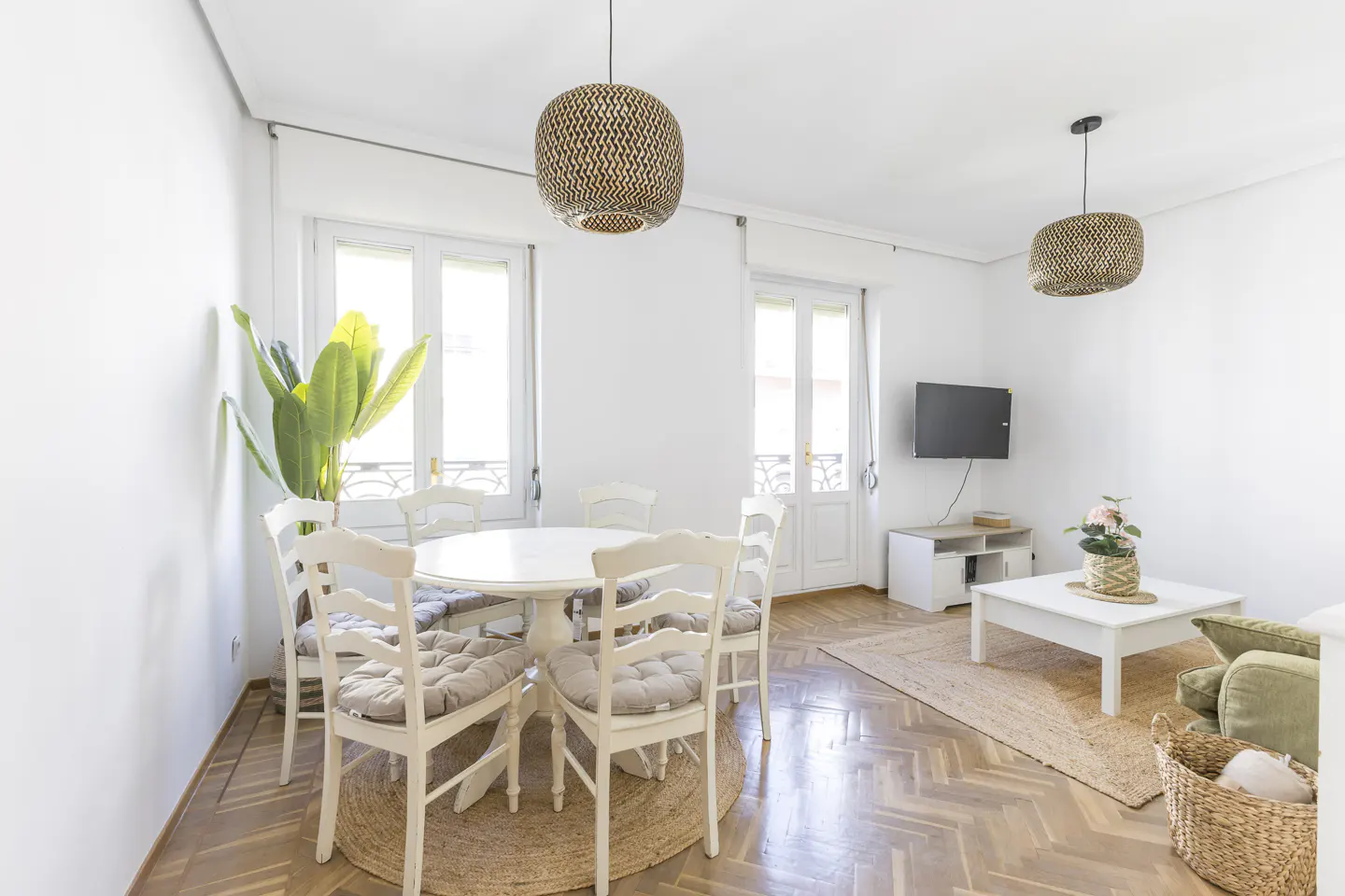 Bright, airy living room with white walls, wood floors, and woven pendant lights. A round white table with six chairs sits near a large plant. A TV and sofa are in the background.