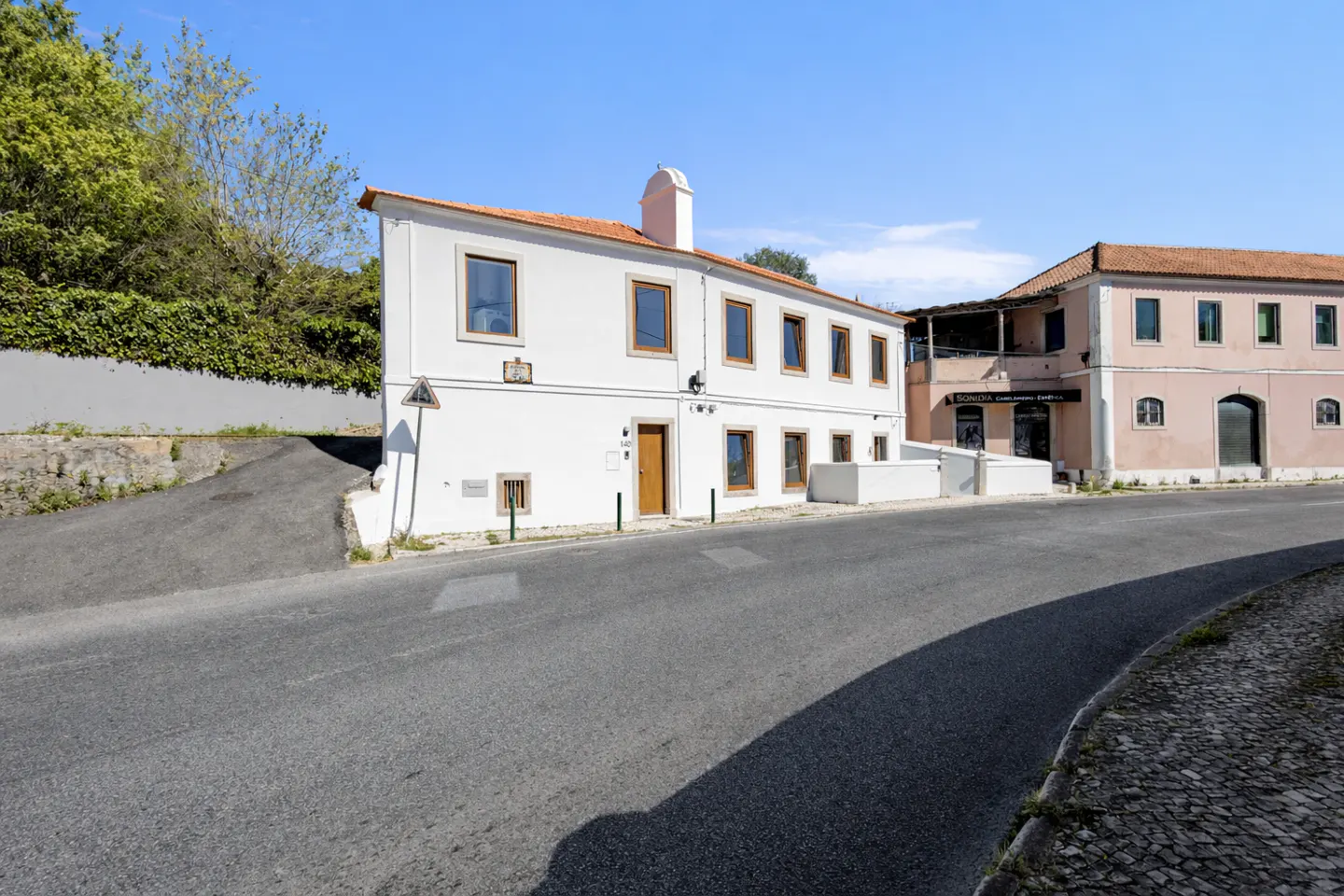 Exterior view of a two-story white building with a brown door and window frames, next to a pink building on a sunny day.
