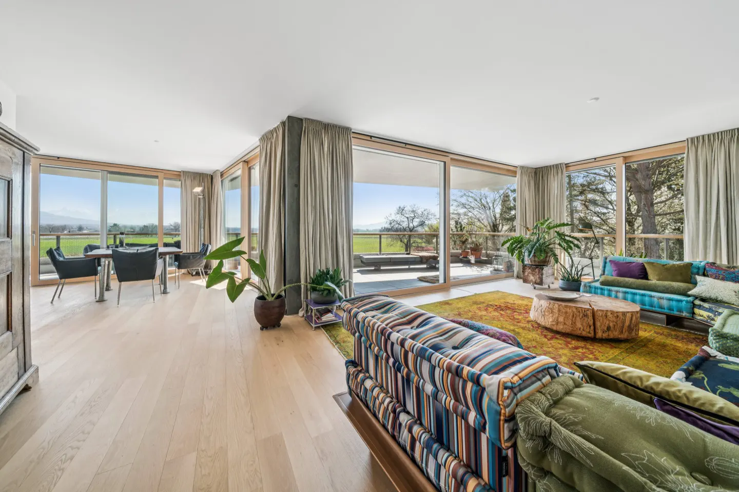 Bright living room with wood floors, large windows, and colorful sofas. Outside, a green field and blue sky are visible.