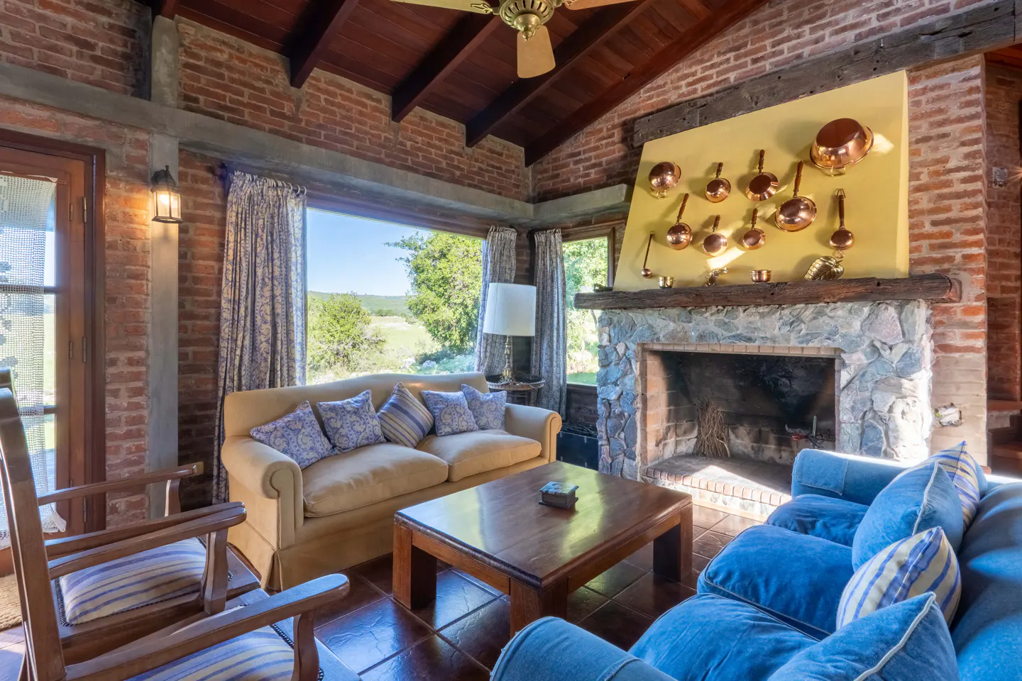 Cozy living room with brick walls, wood ceiling, and stone fireplace. A tan sofa and blue couch face a wooden coffee table. Large windows overlook a green field.