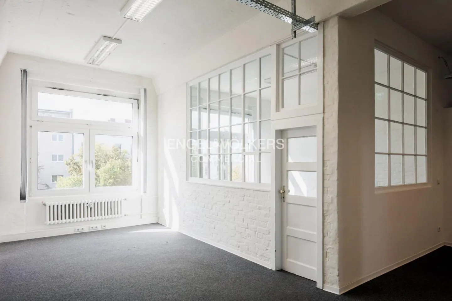 Bright, empty room with gray carpet, white walls, and large windows. A radiator sits below one window, and a white door is on the right.