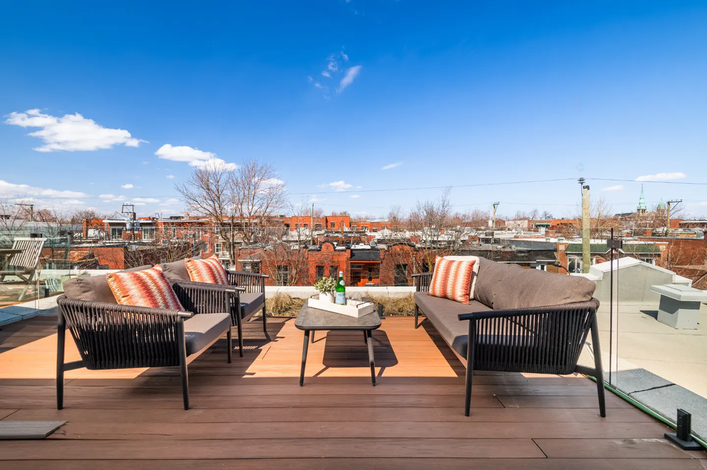 Rooftop patio with outdoor seating. Two black sofas with orange pillows face two chairs. A small table holds drinks. Red brick buildings in the background.