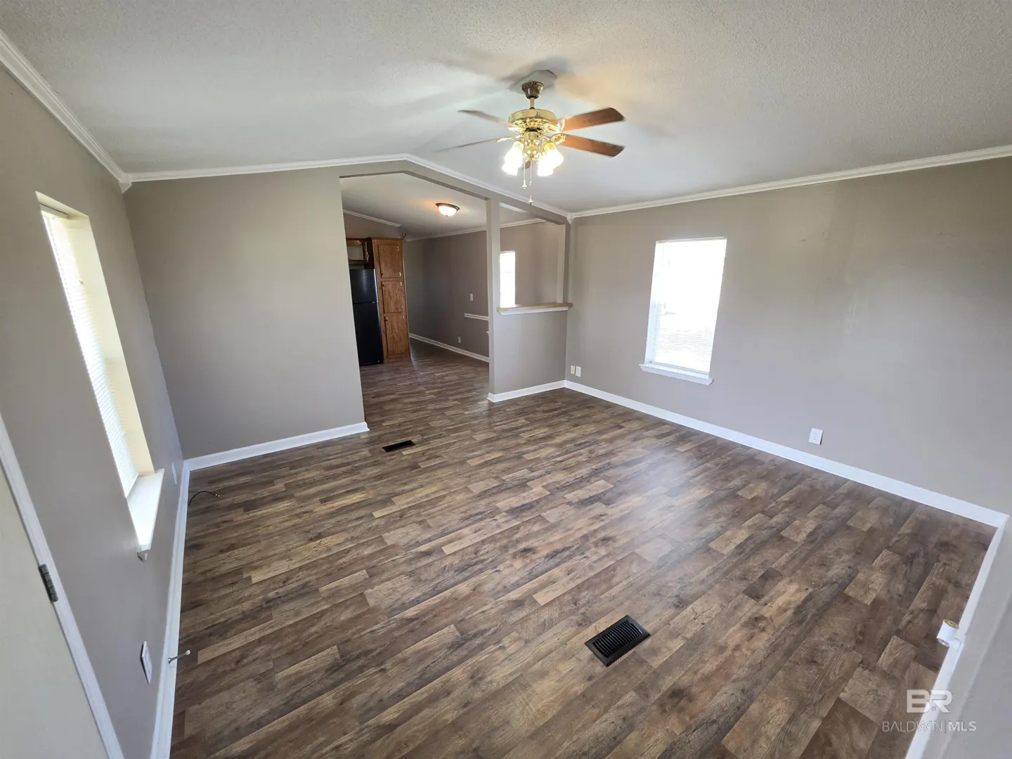 A living room with wood-look floors, gray walls, and white trim. A ceiling fan hangs above, and a partial wall separates the kitchen.