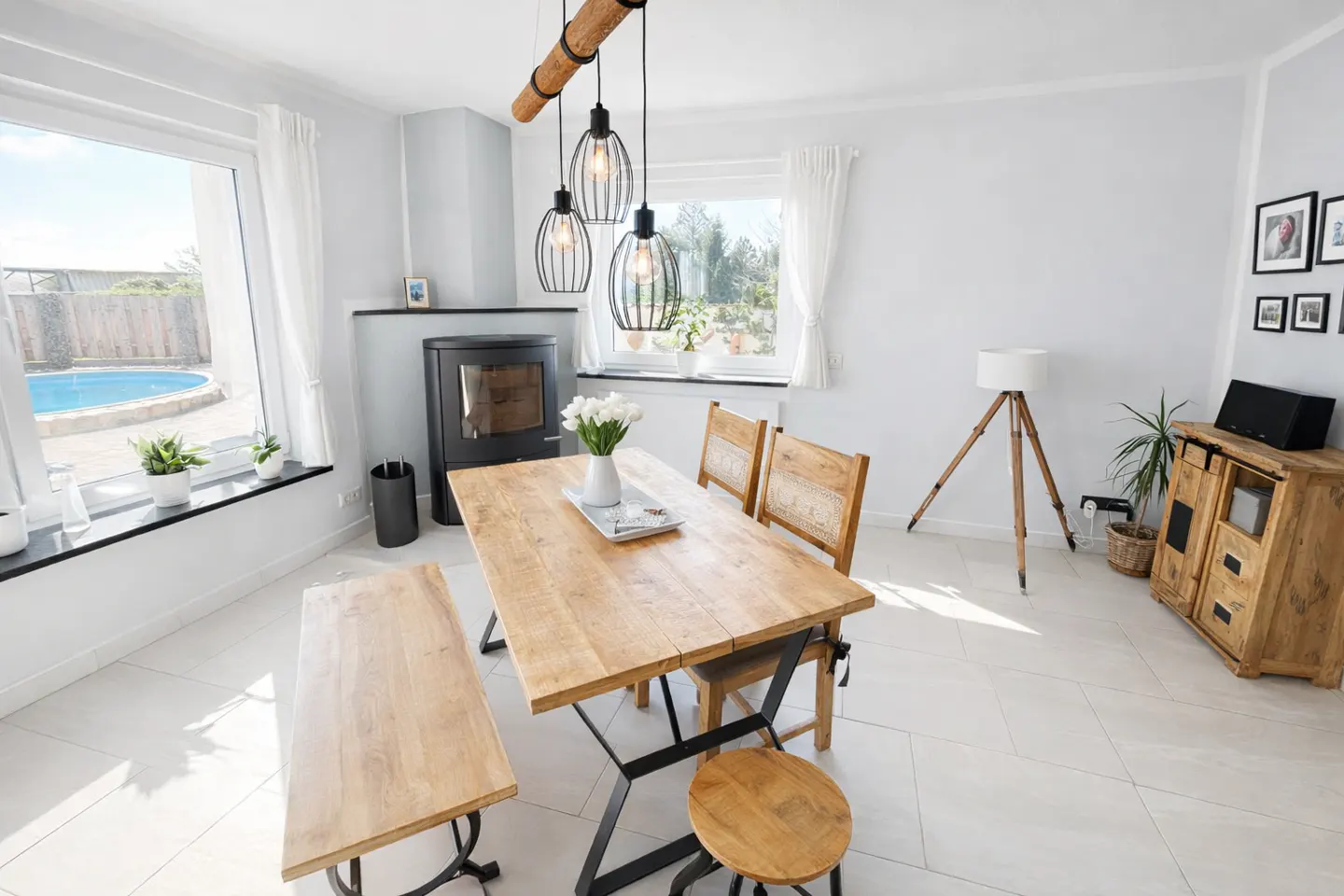 Bright dining room with a wooden table, chairs, and bench. A black fireplace and a view of a pool are visible.