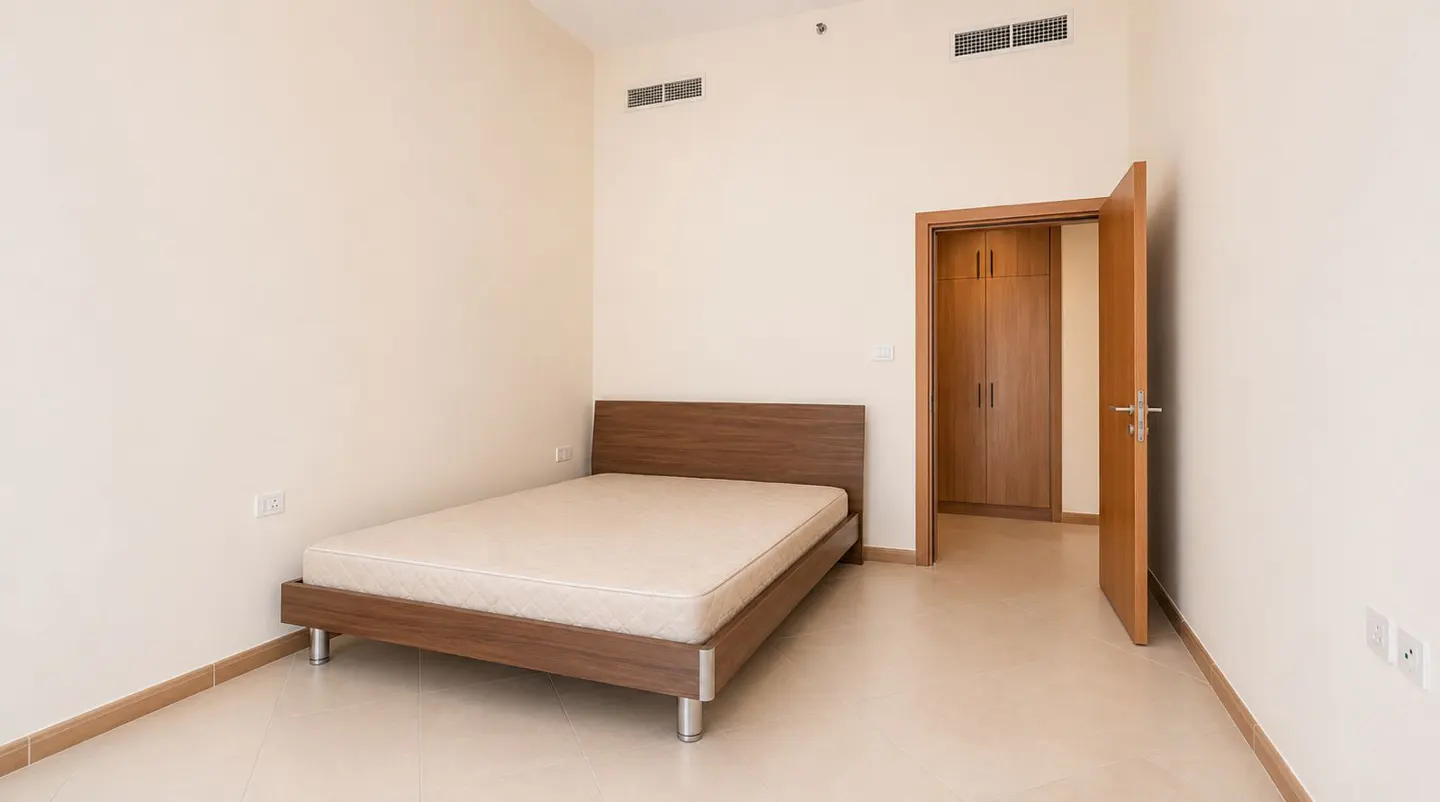 A bedroom with a wooden bed frame, a beige mattress, and an open wooden door leading to a closet. The walls are light beige.