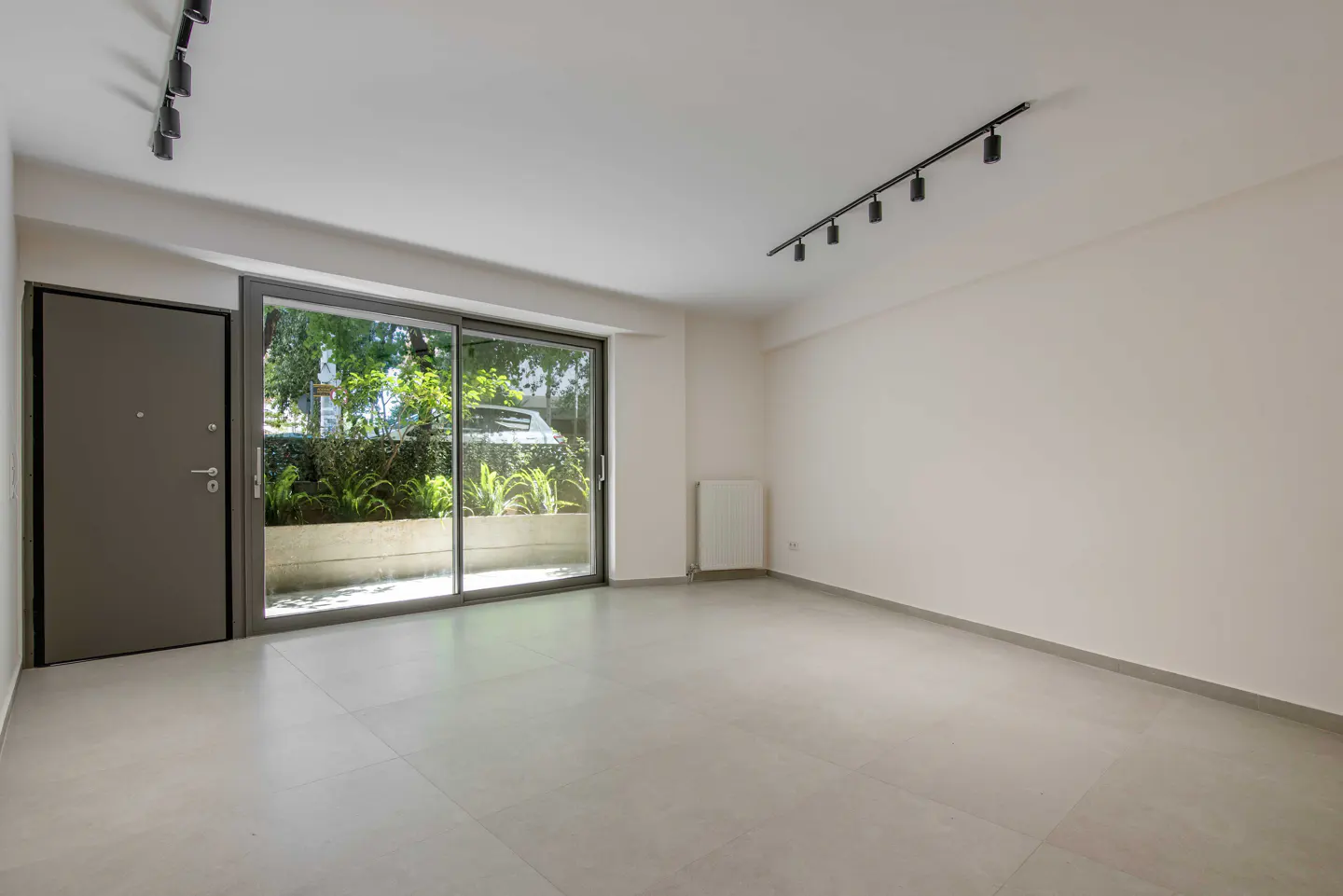 Empty room with white walls, light tile floors, black track lighting, and a sliding glass door looking out to greenery.