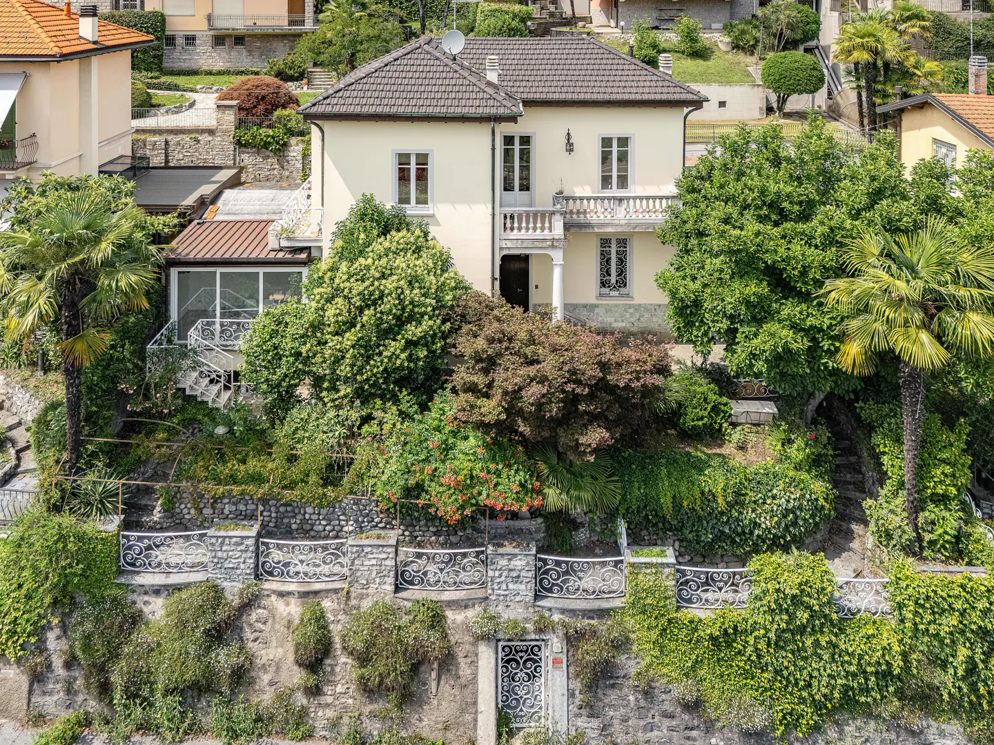 A cream-colored two-story villa with a brown roof, surrounded by lush trees, palm plants, and a stone retaining wall.