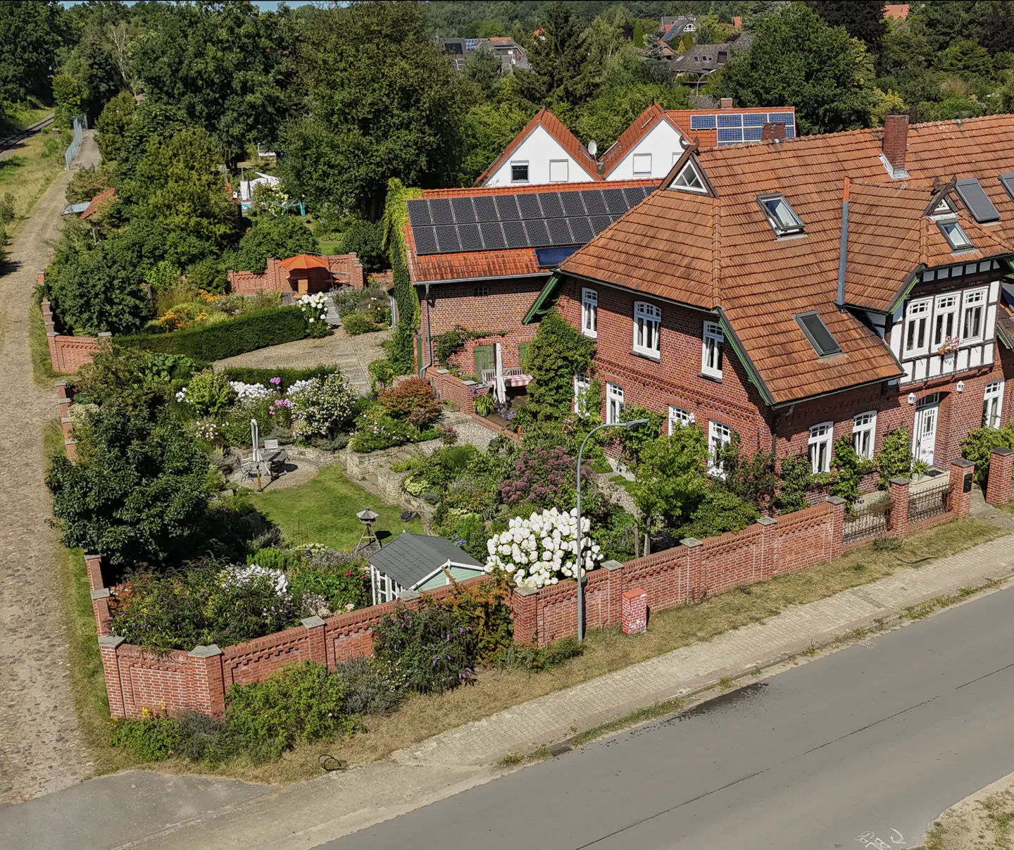 Aerial view of a red brick house with a red tile roof, white trim, and solar panels, surrounded by a lush garden and brick wall.
