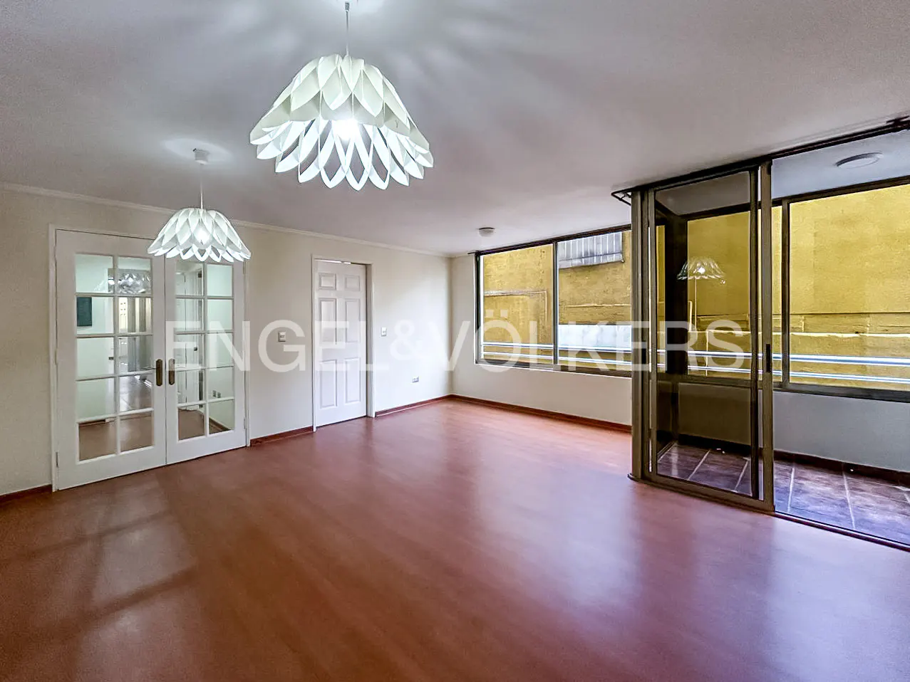 Bright, empty room with wood floors, white walls, and two modern white pendant lights. Large windows and glass doors lead to a balcony.