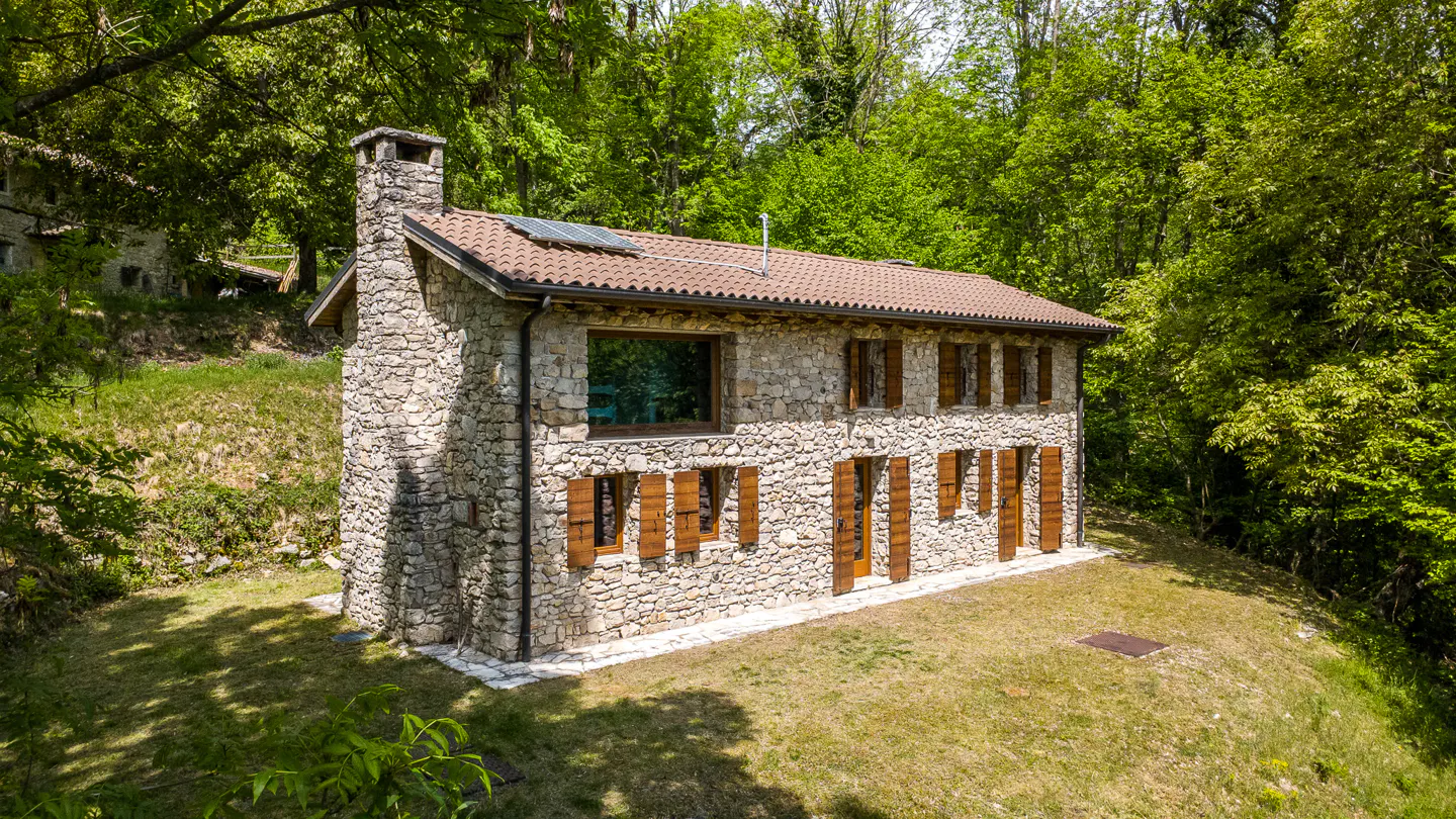 Stone house with brown shutters and a chimney, surrounded by green trees and grass.