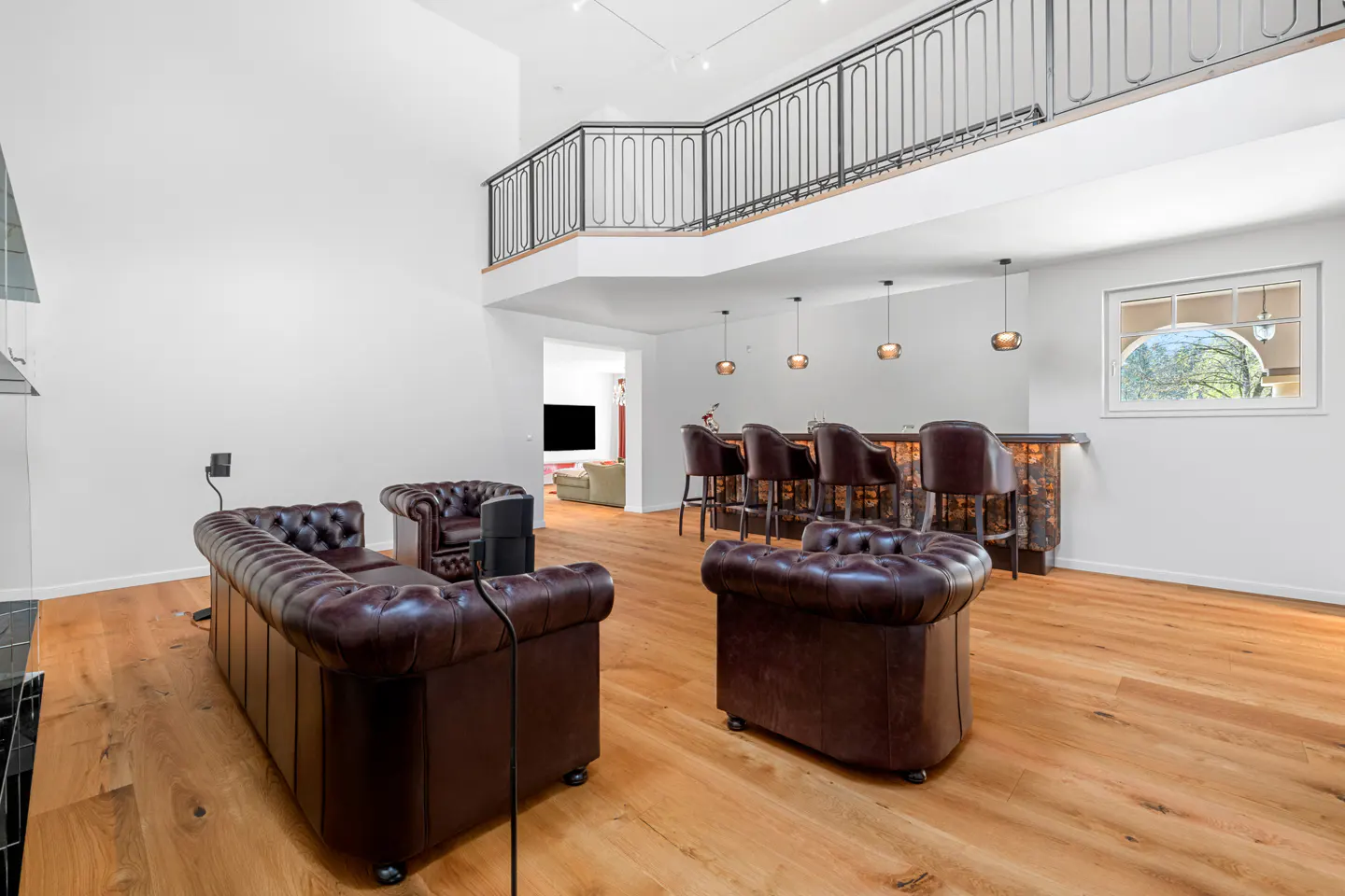 A modern living room with brown leather sofas, a bar with stools, and a second-floor balcony.
