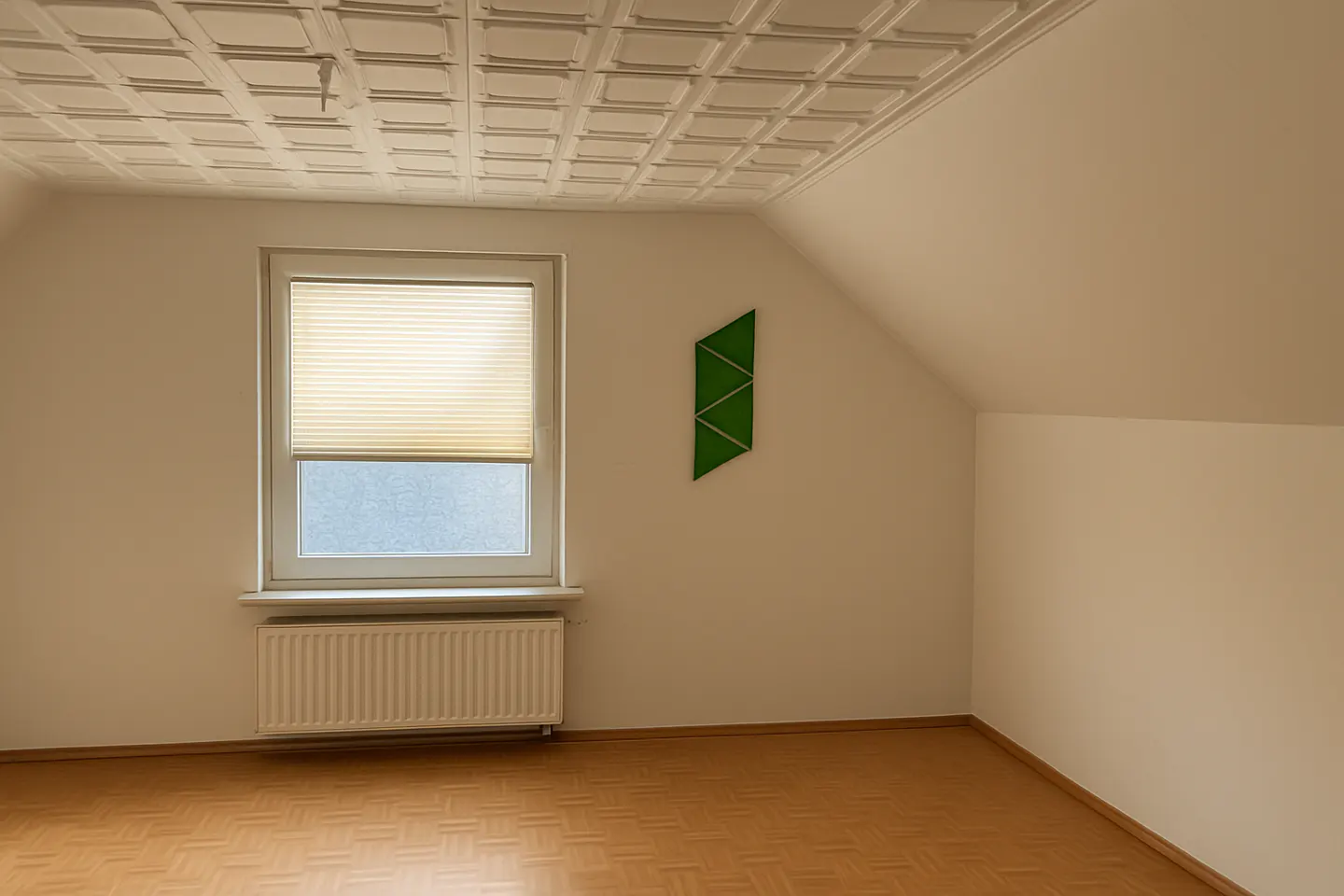 Bright, empty room with parquet floor, white walls, and a textured ceiling. A window with blinds is to the left, and a green geometric wall decoration is to the right.