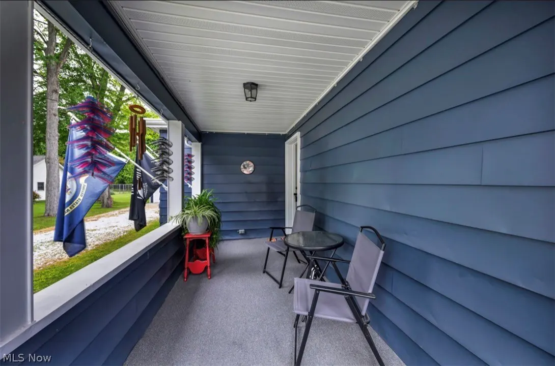 Covered porch with blue siding, white trim, and gray carpet. A small table and chairs sit near a red plant stand. Flags and wind chimes hang near the porch opening.