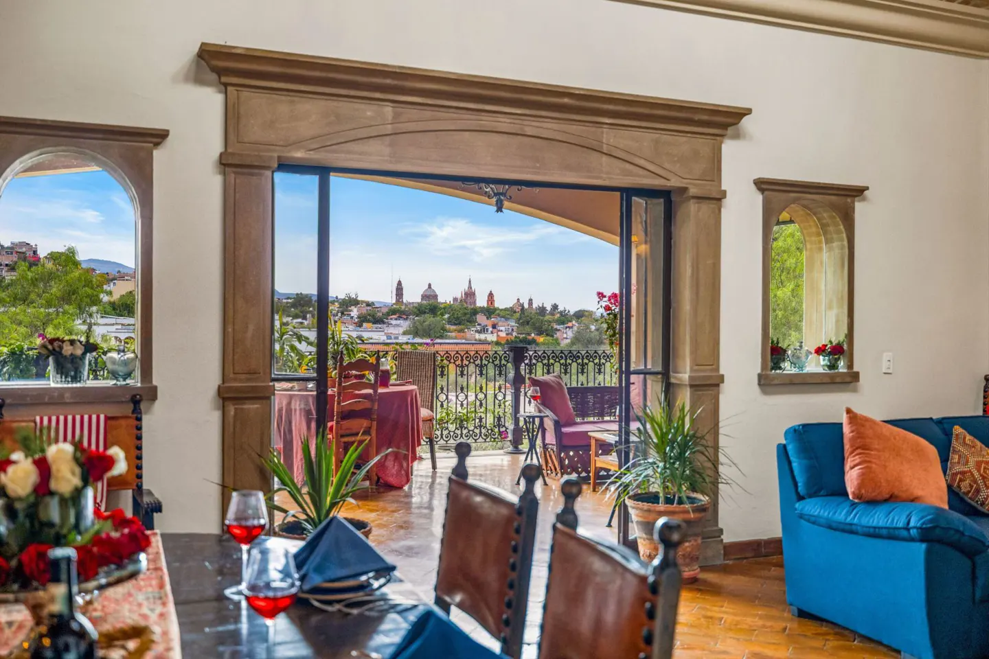 Interior view of a dining room with a balcony overlooking a city skyline under a blue sky. The room has a blue sofa and a wooden table.