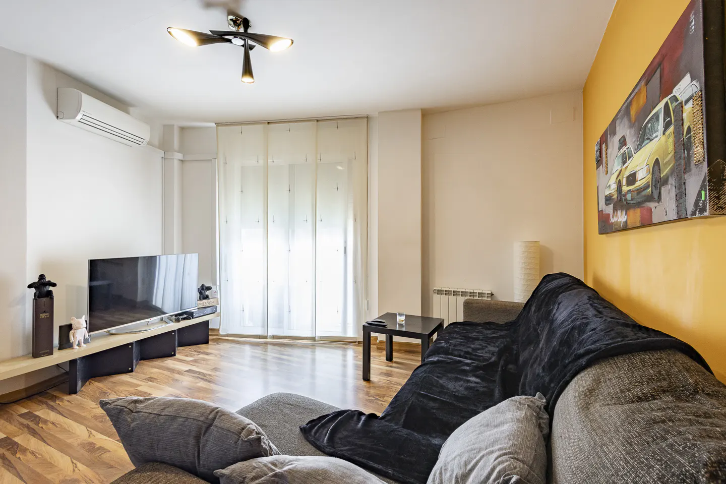 Living room with gray sectional sofa, black throw, TV, and yellow accent wall with car art. Hardwood floors and sheer curtains.