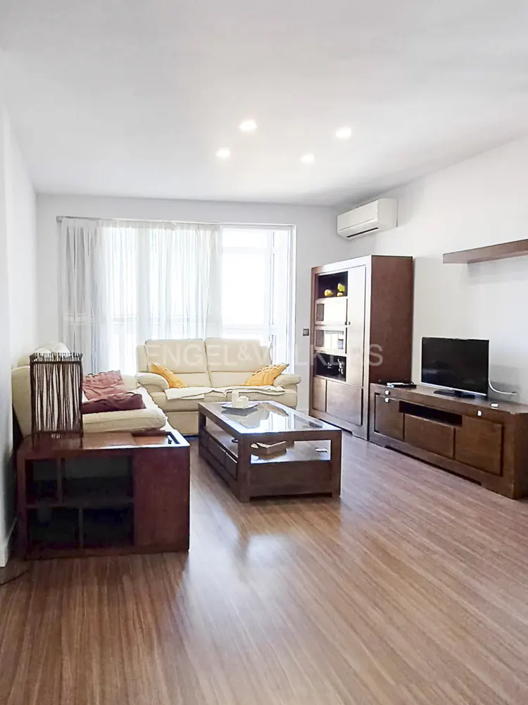 Bright living room with wood floors, white sofa, and dark wood furniture. Large windows with sheer curtains let in natural light.
