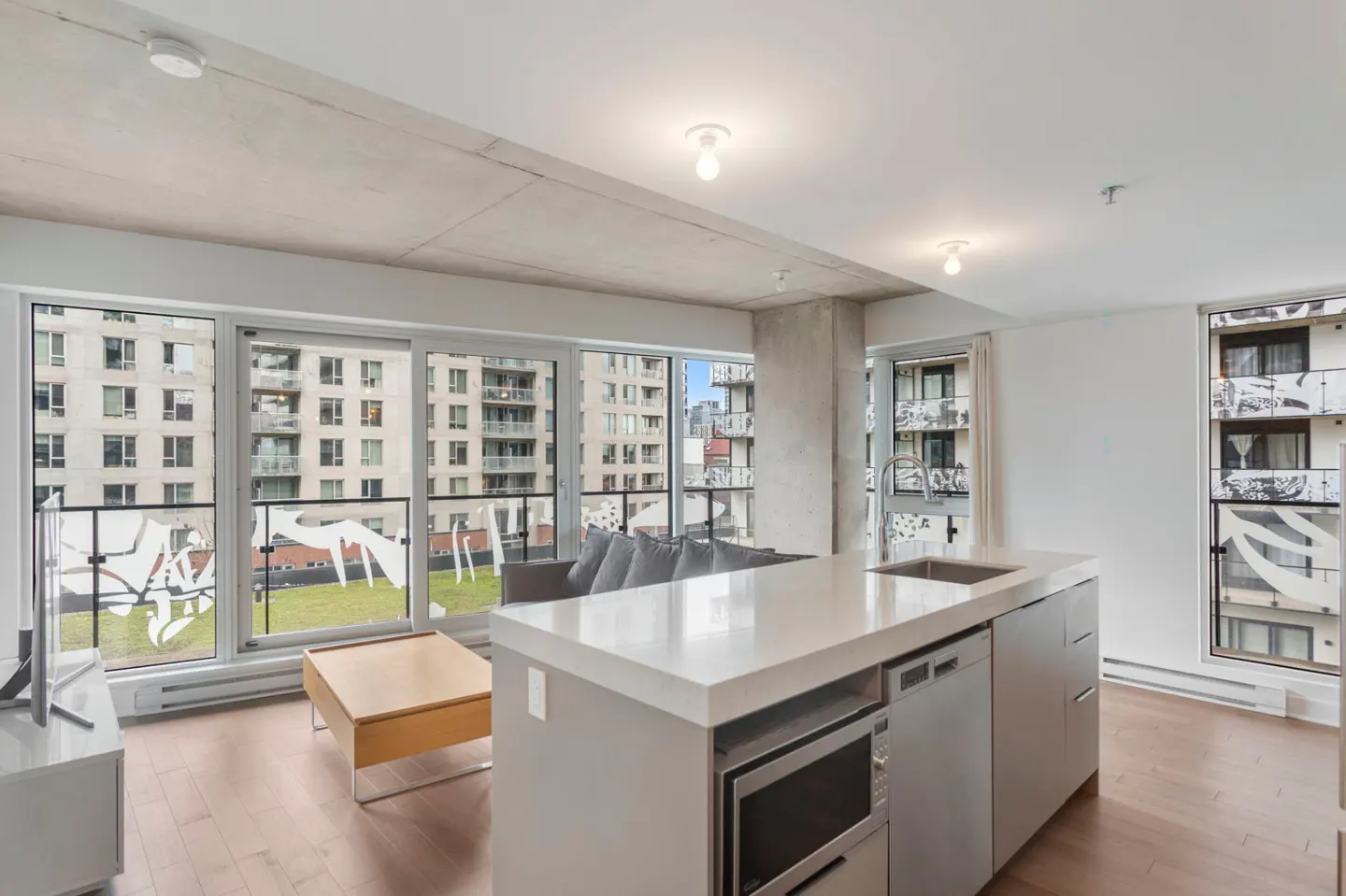 Modern apartment with a kitchen island, light wood floors, and floor-to-ceiling windows overlooking a city courtyard.