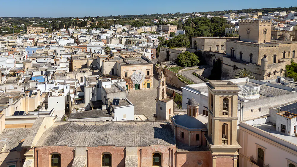 Aerial view of Castro, Italy, featuring a pink church, bell tower, and a castle in the background. The town is densely packed with white buildings.