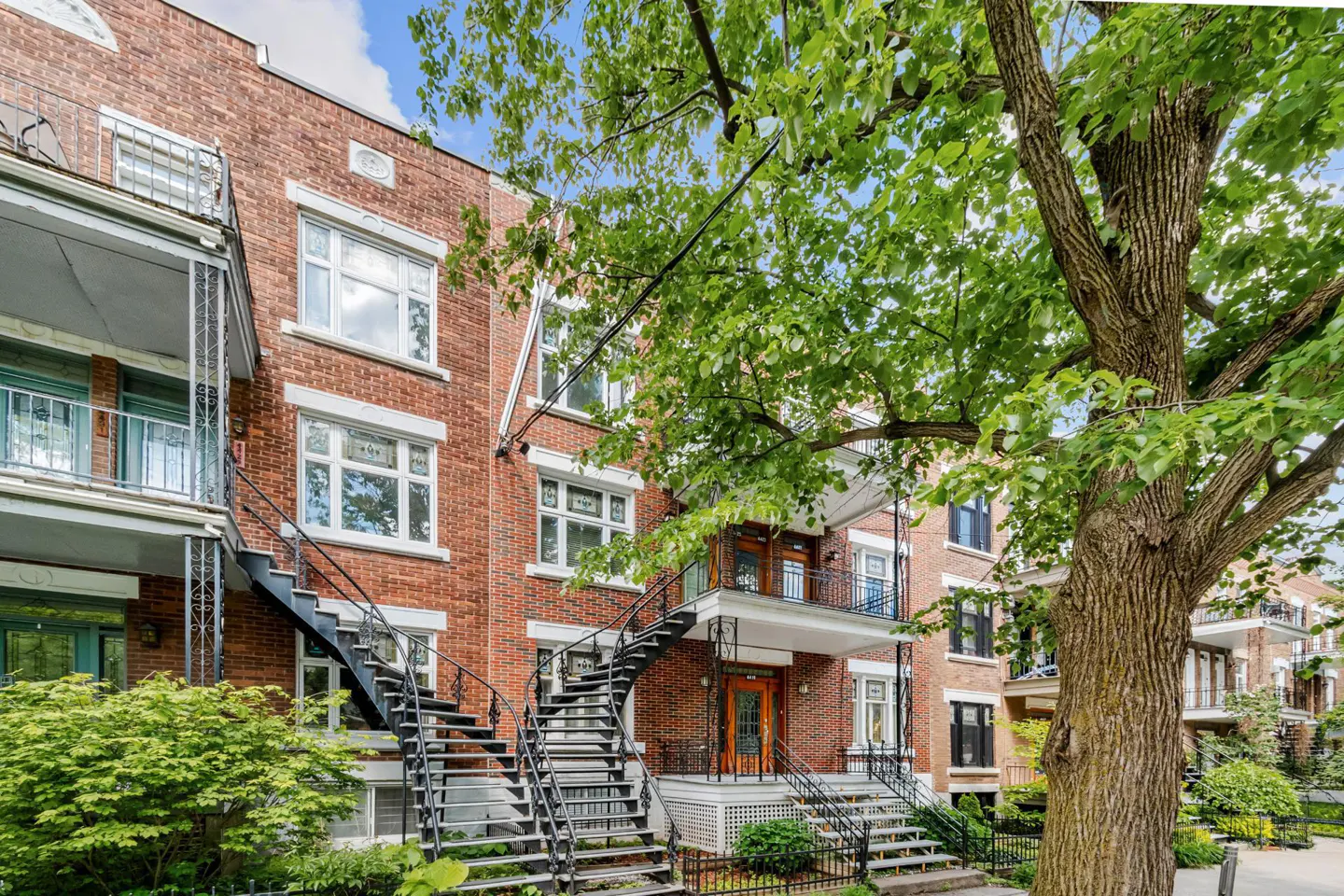 A three-story red brick apartment building with black metal exterior staircases and a large tree in the foreground.