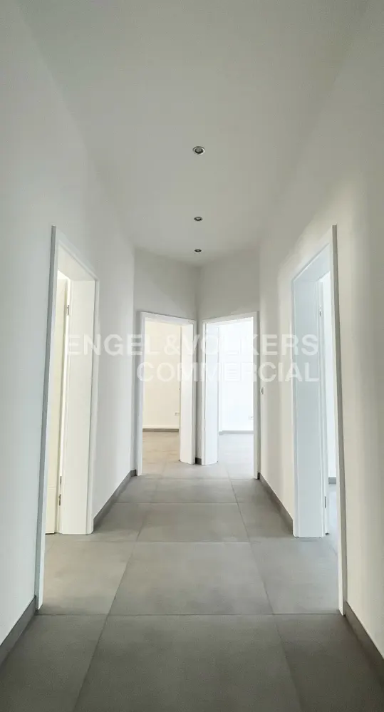 A bright hallway with white walls, gray tile floors, and open doorways leading to other rooms. Recessed lighting is visible in the ceiling.