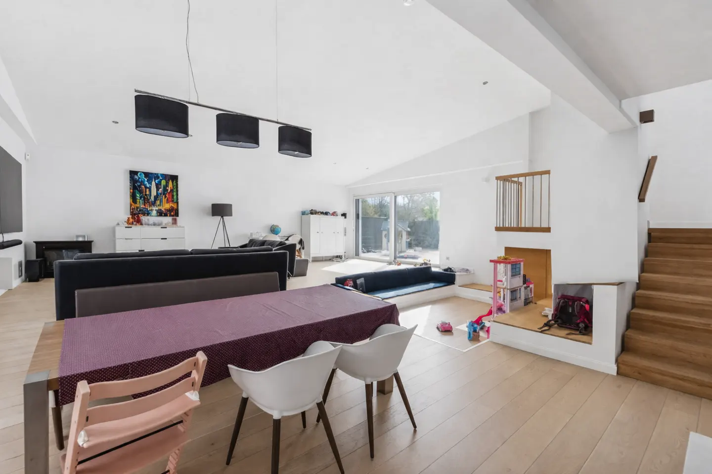 Bright, open-plan living space with white walls, wood floors, dining table, sofa, and stairs. Natural light floods in through sliding glass doors.