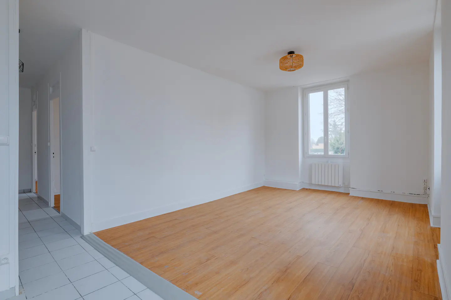 Bright, empty room with white walls, wood floors, and a window. A hallway with tile floors is visible to the left.