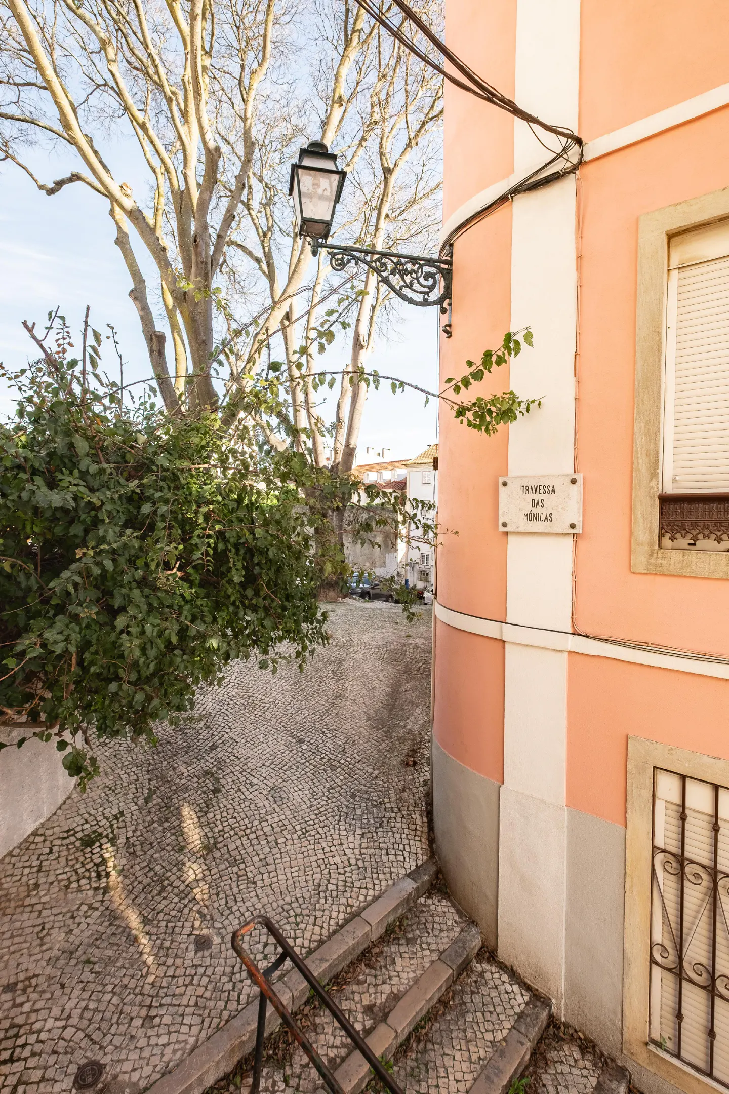 Cobblestone street view with steps and a metal handrail leading down. A pink building with a street sign is on the right.