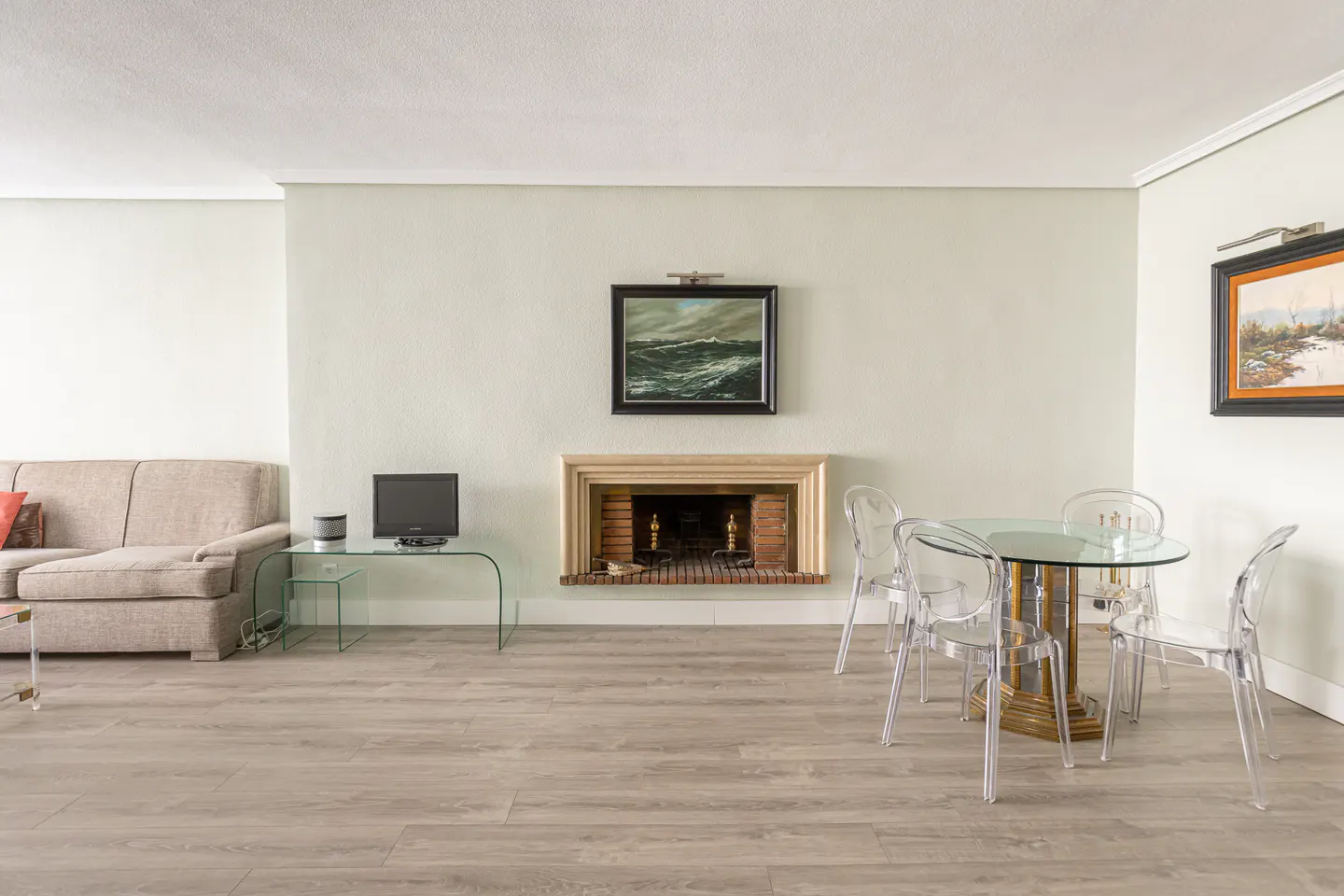 Living room with beige sofa, glass TV stand, fireplace with art above, and a glass dining table with clear chairs.