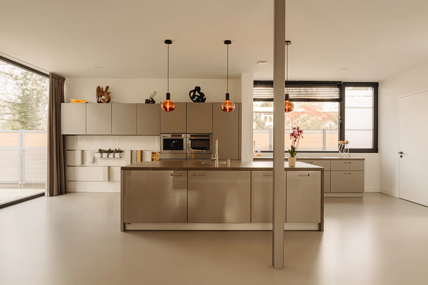 Modern kitchen with gray cabinets, island, and orange pendant lights. Large windows provide natural light. Minimalist decor.