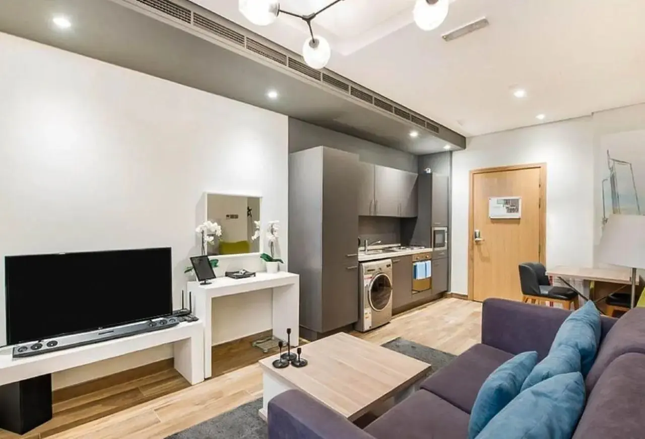 A modern apartment interior with a purple sofa, TV, white console table, and a compact kitchen with gray cabinets.
