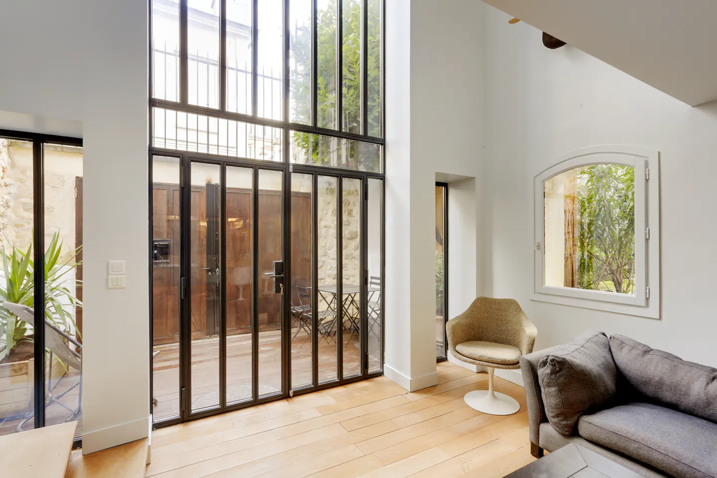 Bright living room with white walls, wood floors, and black framed windows. A gray couch and tan chair sit near a window overlooking greenery.