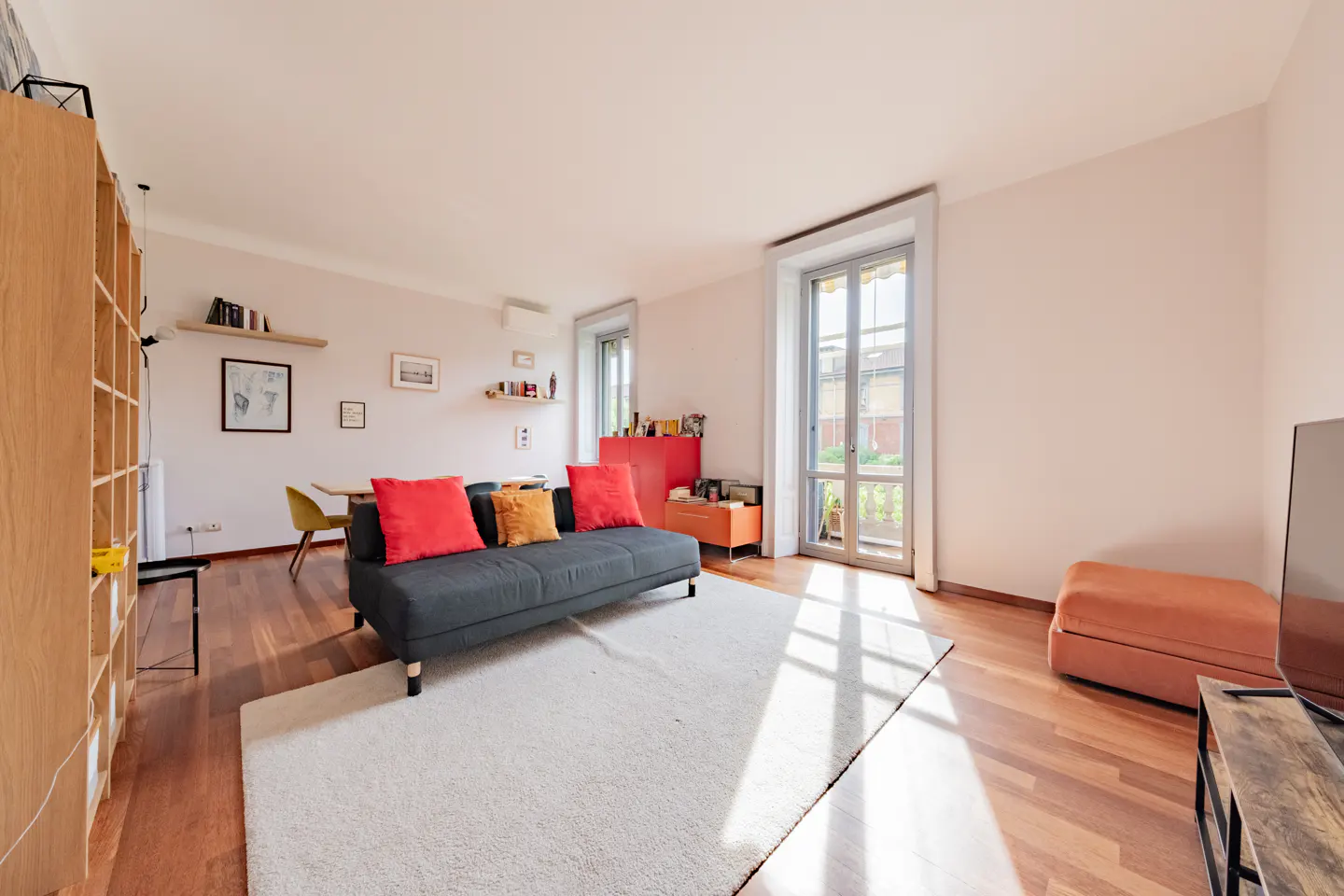Bright living room with a gray sofa, red and gold pillows, white rug, and wood floors. A bookcase and dining table are visible.