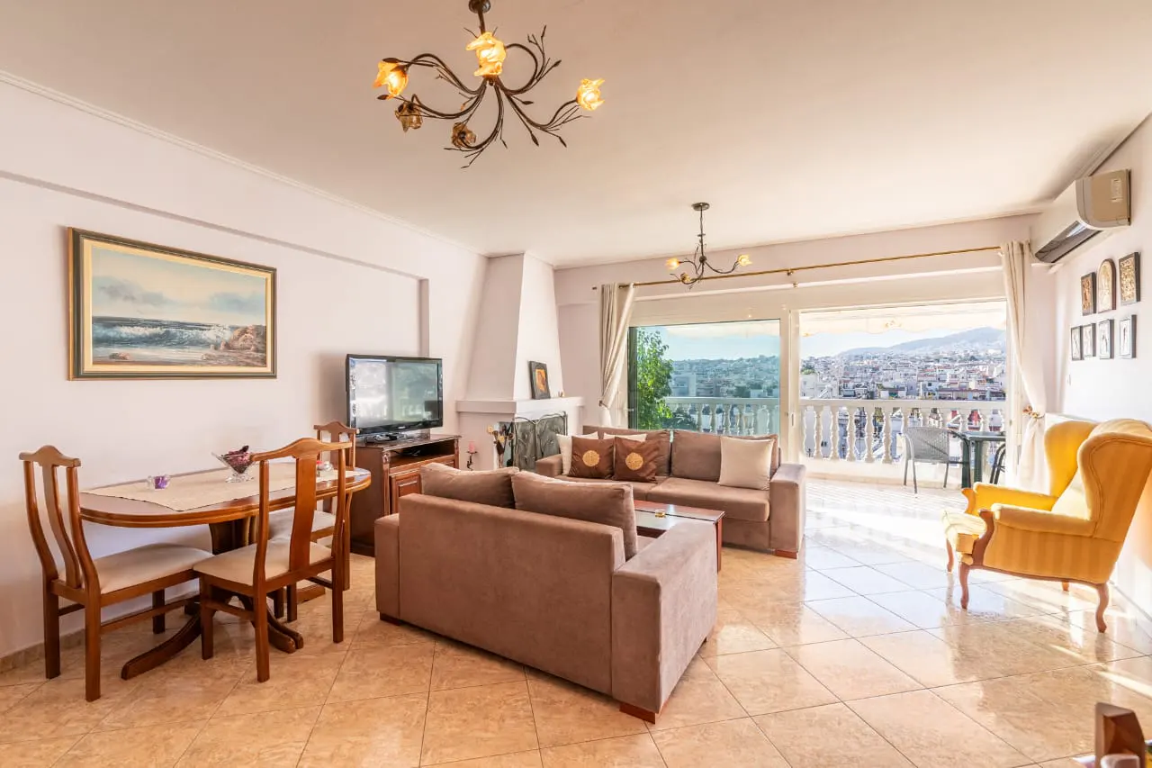 Bright living room with brown sofas, yellow armchair, and dining table. Balcony with city view. Tiled floor and decorative lighting.
