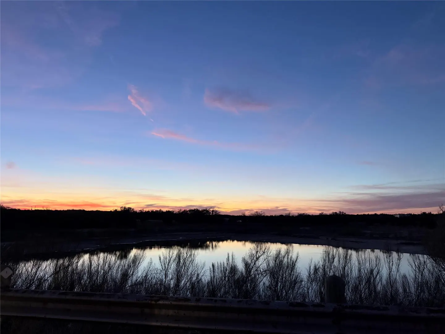 A serene sunset over a calm lake, with silhouettes of trees and a guardrail in the foreground against a colorful sky.
