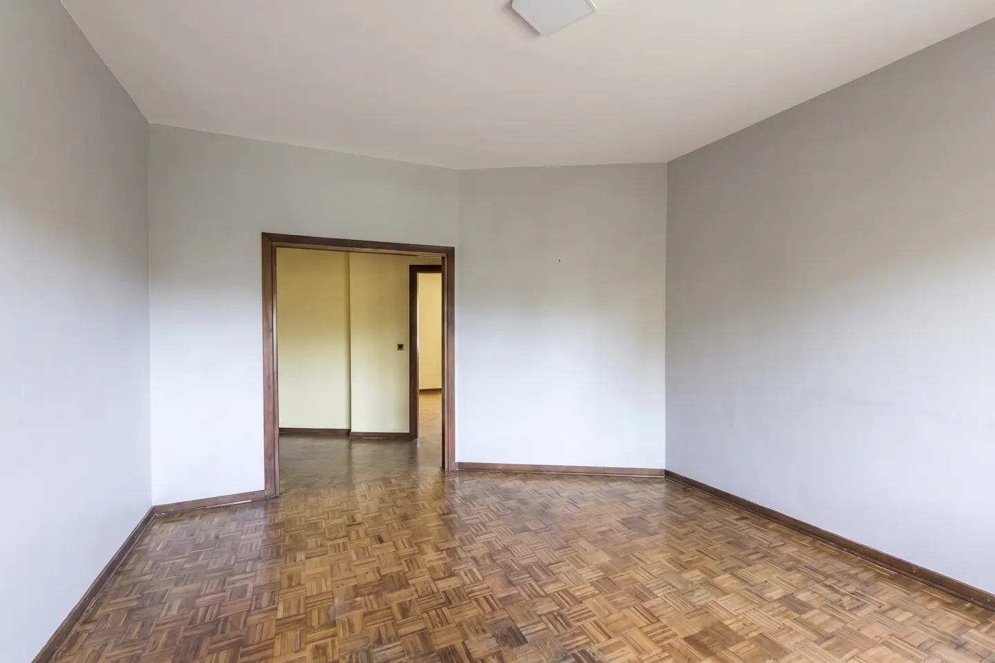Empty room with light gray walls, parquet wood flooring, and a doorway leading to a yellow-walled hallway.
