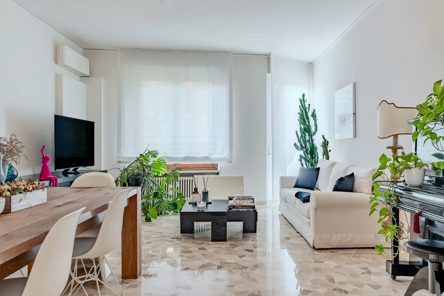 Bright living room with white walls, a wood table, white chairs, and a white sofa with plants.