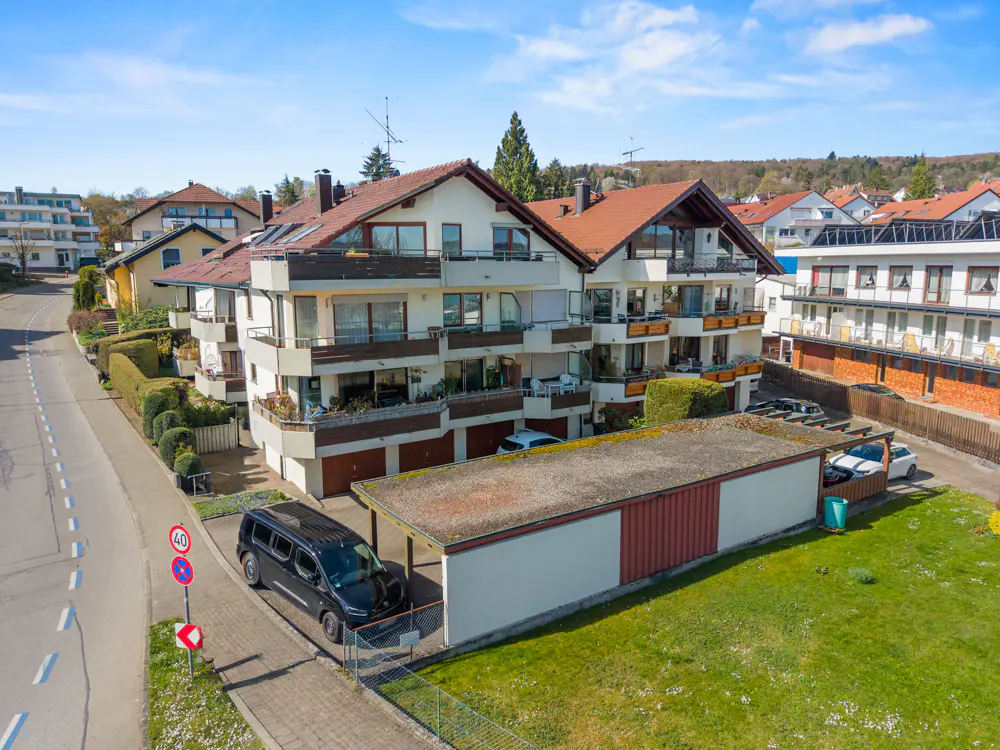Three-story apartment building with balconies and red-tiled roof, surrounded by green lawns and other houses.