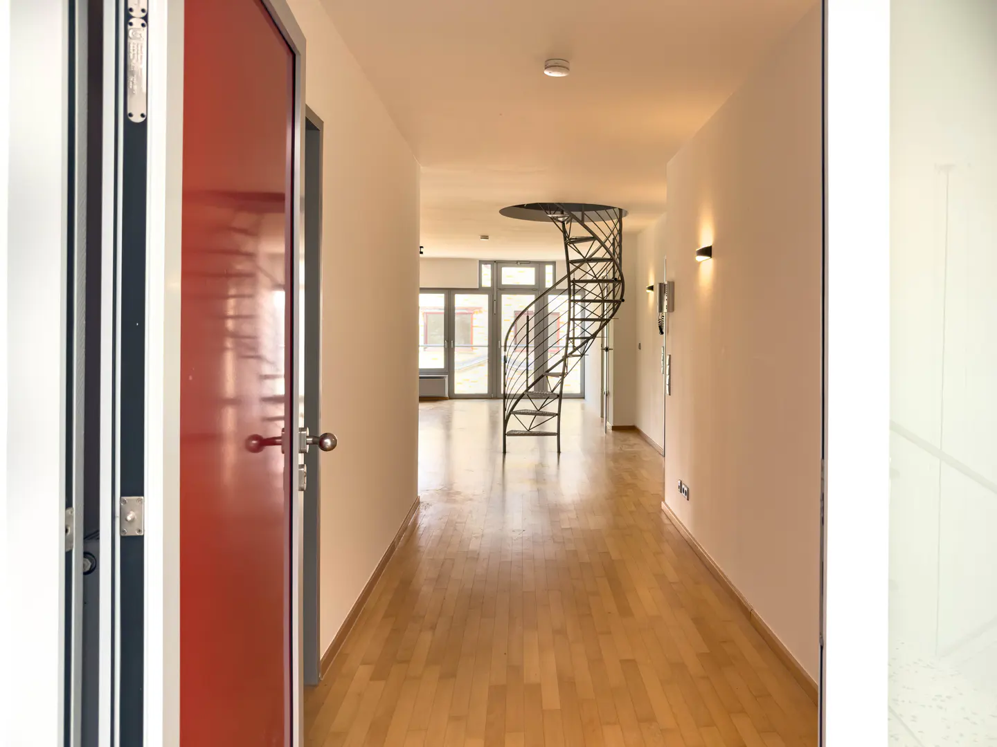 Hallway view with a red door on the left, light wood floors, and a black spiral staircase in the background.