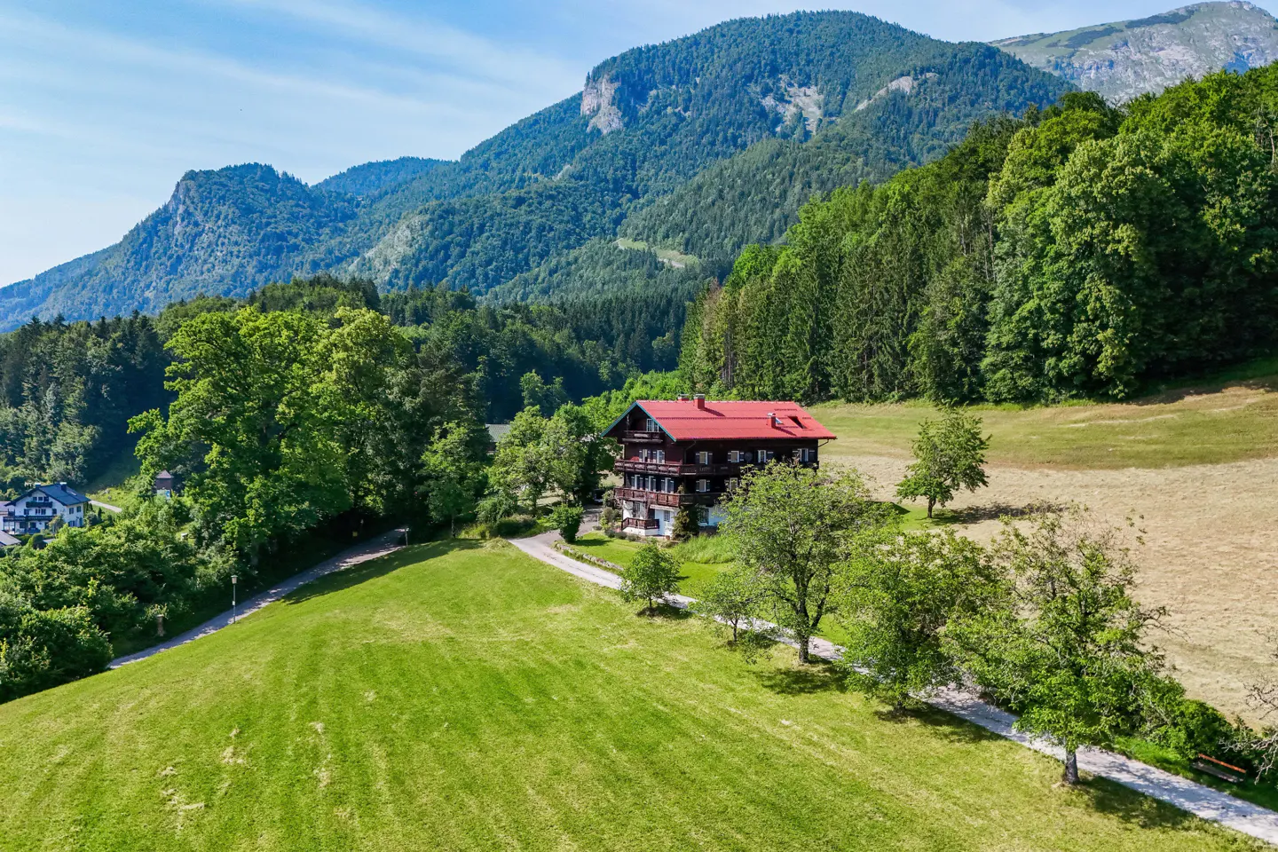 A chalet with a red roof sits on a green hillside, backed by mountains and trees under a blue sky.