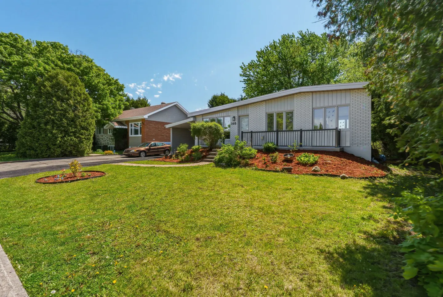 Single-story white brick house with a front porch, large green lawn, and landscaped garden beds under a clear blue sky.