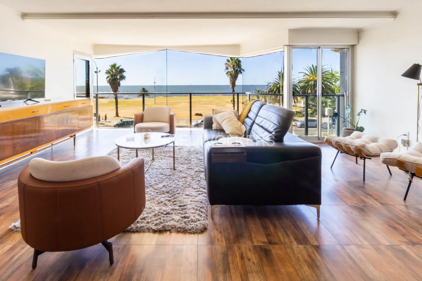 Bright living room with a black leather sofa, chairs, and a view of the beach through large windows.