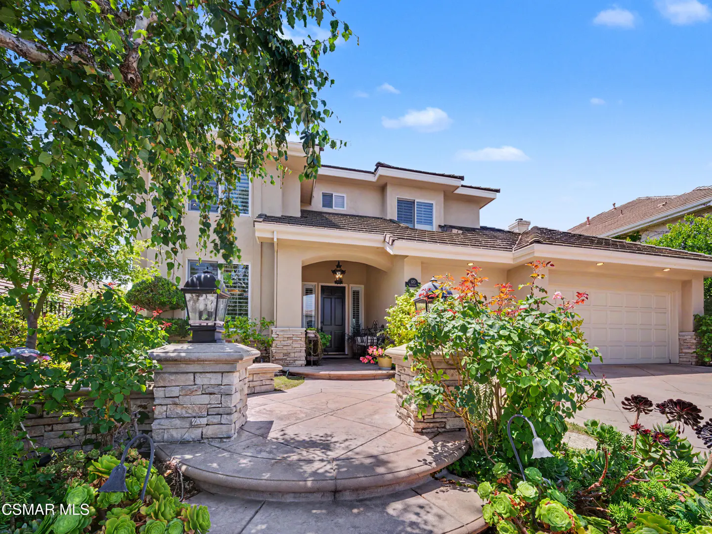 Two-story beige suburban home with a stone-accented entryway, curved concrete path, and lush front garden under blue sky.