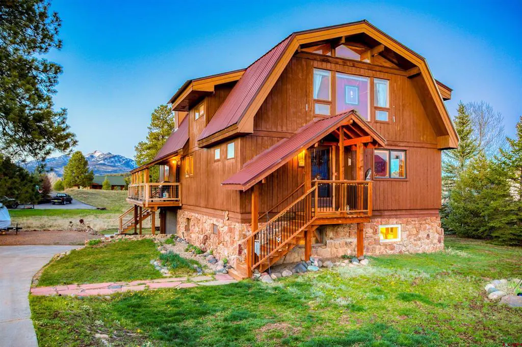 A two-story brown house with a red roof, stone foundation, and green lawn. Trees and mountains are in the background.