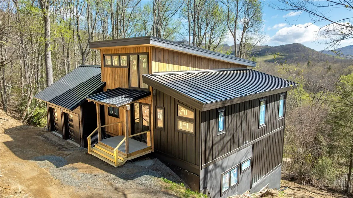 Modern two-story home with black metal roof and wood siding on a wooded lot. Mountains are visible in the background.